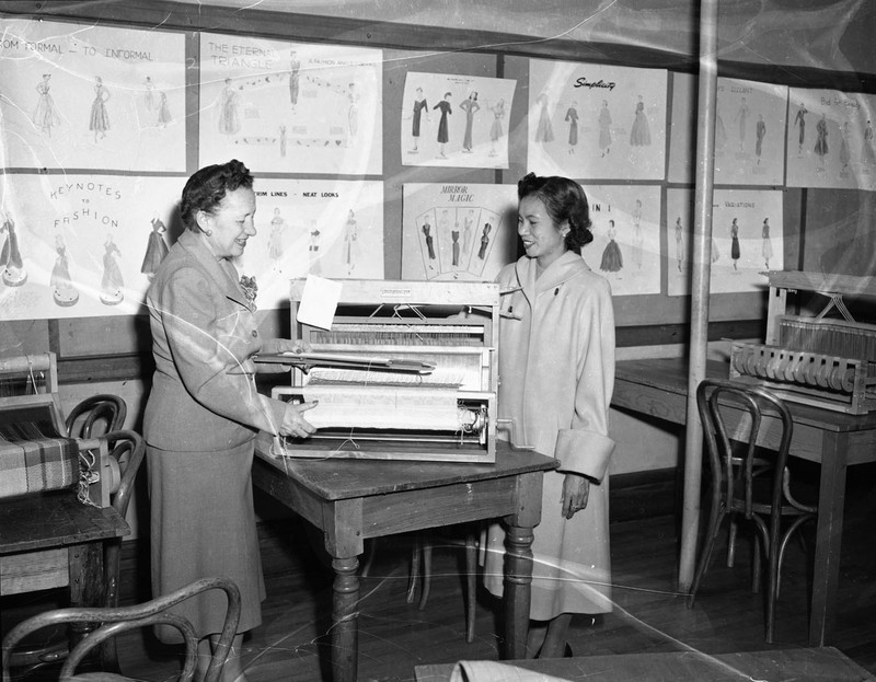 Margret Ritchie (left),Professor of Home Economics at University of Idaho, speaking to foreign student Gertrude Vallejos (right) next to a textile weaving loom. Fashion sewing posters cover the wall behind them.
