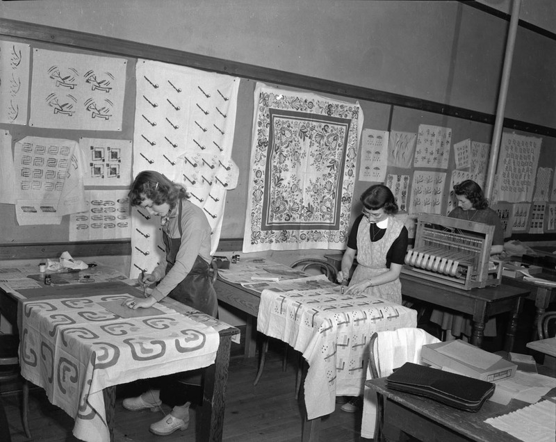 Two women tracing patterns onto cloth. A third woman is operating a weaving loom. Fabrics are pinned to the wall in the background.