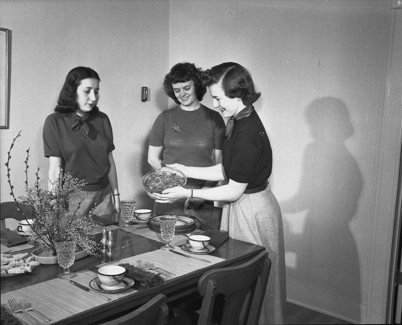 Three women standing around a set dining table, looking at a prepared dish.