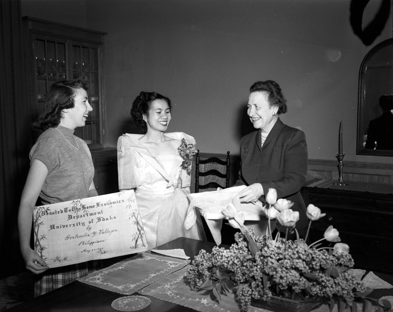 Foreign student from the Philippines, Getrude Vallejos (middle), making a donation to the Department of Home Economics in the company of Margret Ritchie (right), Professor of Home Economics at the University of Idaho, and an unidentified student (left) at a dining table. Vallejos is wearing a white gown and floral boutonniere. A certificate of the donation is in the hands of the woman on the left.