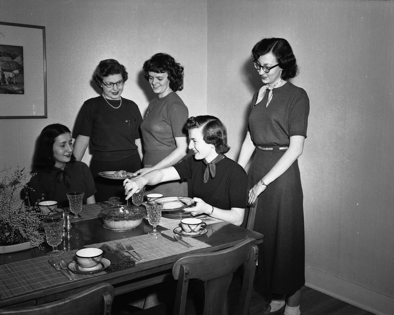 Five women around a set dining table, serving food.