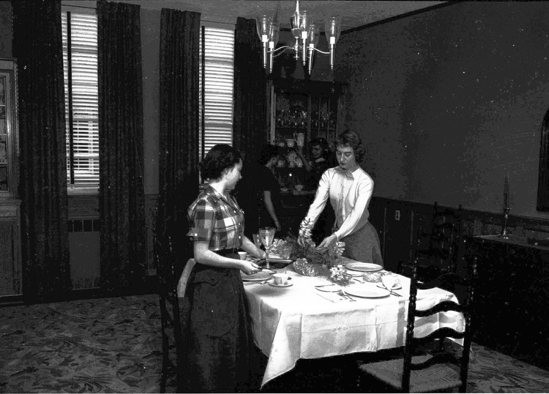 Four women setting a dining table, two setting dishes on the table, and two in the back collecting dishes from a cabinet.