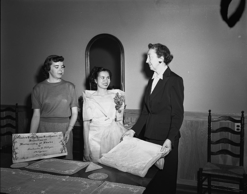 Foreign student from the Philippines, Getrude Vallejos (middle), making a donation to the Department of Home Economics in the company of an unidentified faculty member and an unidentified student (left) at a dining table. Vallejos is wearing a white gown and floral boutonniere. A certificate of the donation is in the hands of the woman on the left.