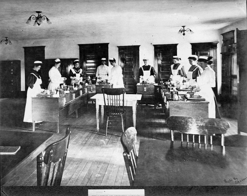 Women dressed in aprons stand behind tables and prepare food on a tray.