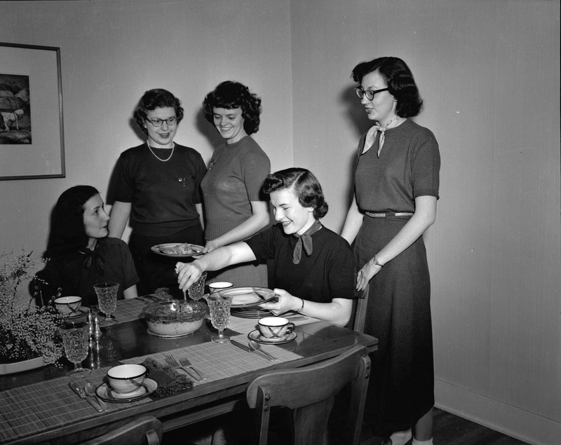 Five Home Economics students around a set dining table, serving a dish of food.