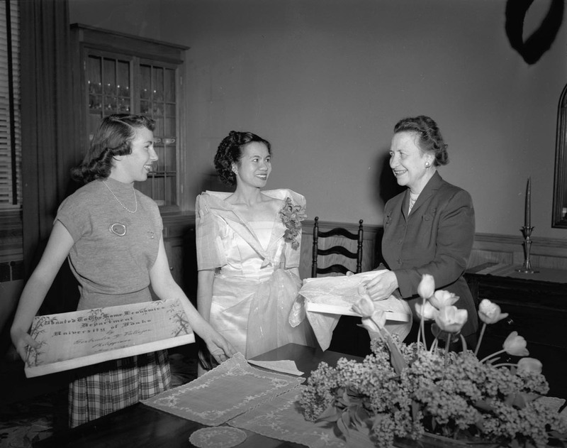 Foreign student from the Philippines, Getrude Vallejos (middle), making a donation to the Department of Home Economics in the company of Margret Ritchie (right), Professor of Home Economics at the University of Idaho, and an unidentified student (left) at a dining table. Vallejos is wearing a white gown and floral boutonniere. A certificate of the donation is in the hands of the woman on the left.