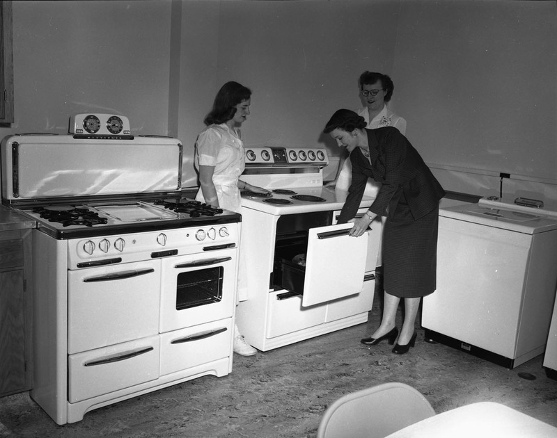 Three women standing around an oven, one opening it.