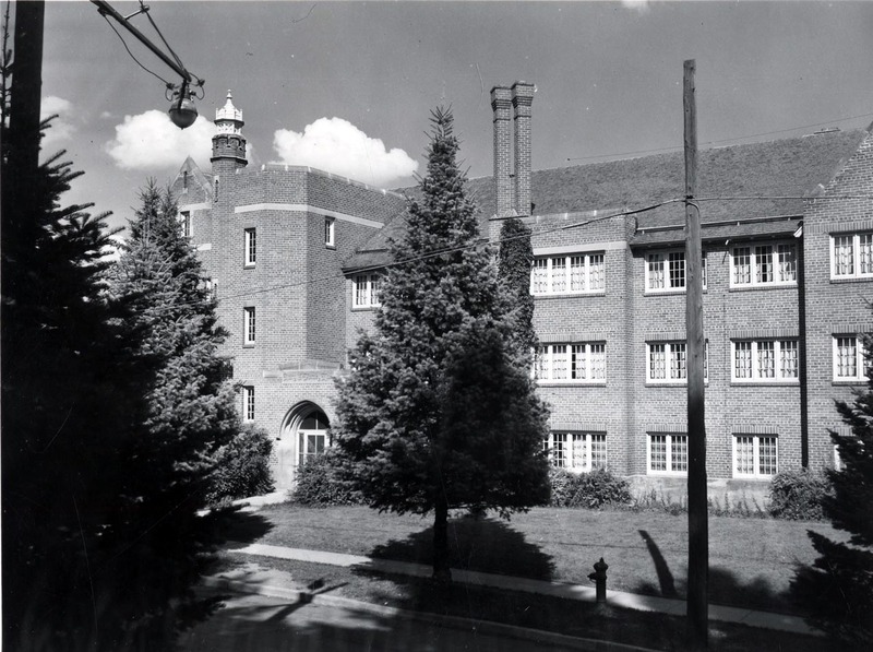 A photograph of Forney Hall, a three story brick and concrete building, built in 1927 as a women's dormitory. Trees block some view of the building in the photograph.