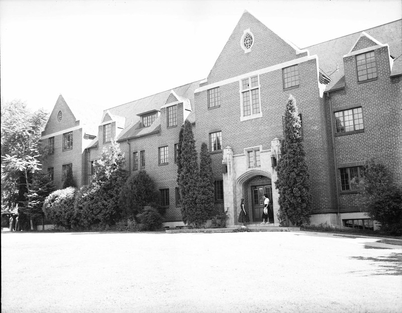 A photograph of the front of Forney Hall, a three story brick and concrete building. Built in 1927 as a women's dormitory. Two women can be seen standing in front of the doors to the building.