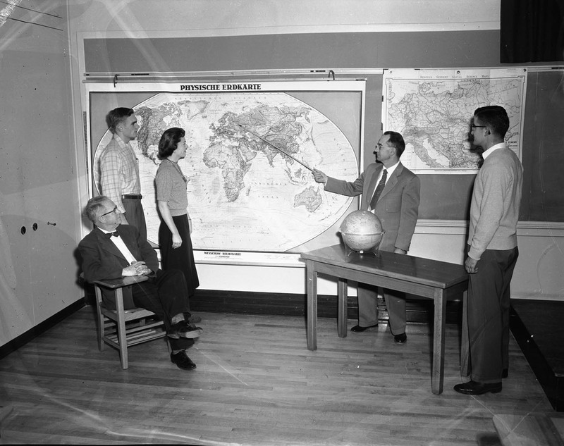 A professor of geography pointing to Moscow, Russia on a German world map in a classroom. Students are on either side of the map. A globe sits on the desk to the right.