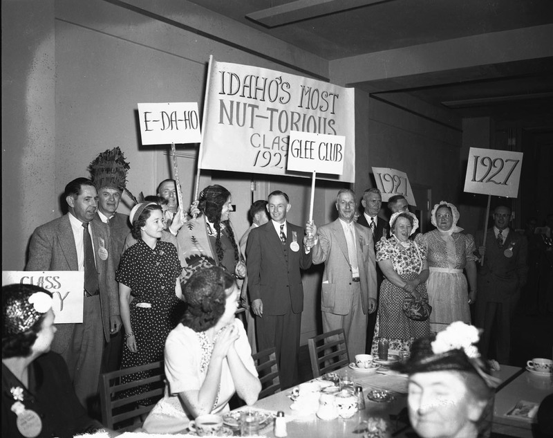 A group of period-costumed University of Idaho graduates holding class of 1927 signs. People dining can be seen in the foreground.