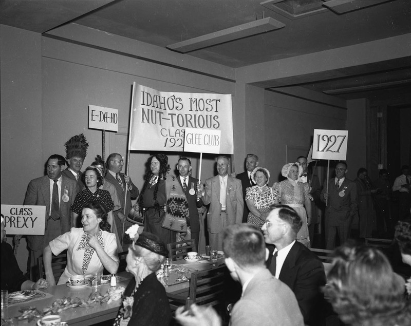 A group of period-costumed University of Idaho graduates holding class of 1927 signs. People dining can be seen in the foreground.