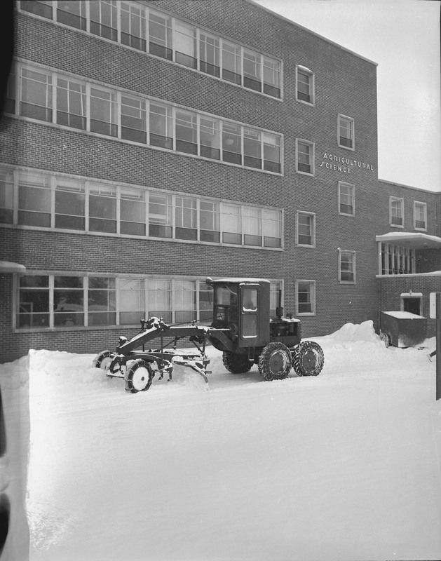 A picture of farming equipment equipped with a snow plow outside of the Agricultural Science Building. Snow can be seen on the ground.