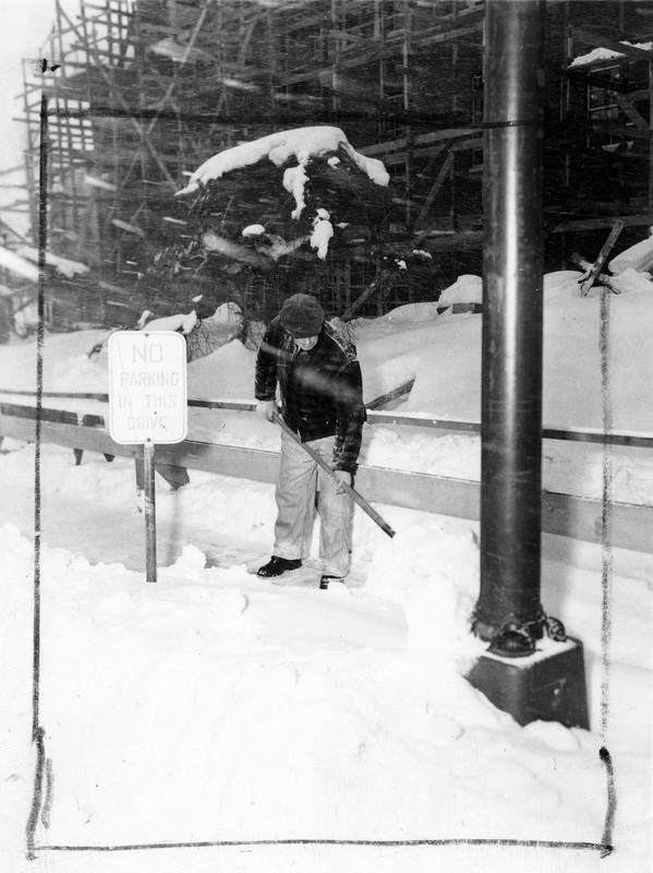 A man shoveling snow next to a sign that reads 'No Parking in this Drive'. A building under construction can be seen in the background.