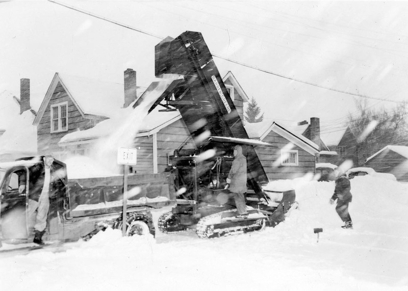 A snow excavator pouring snow into a dump truck during a snowstorm, three men can be seen operating the truck and excavator.