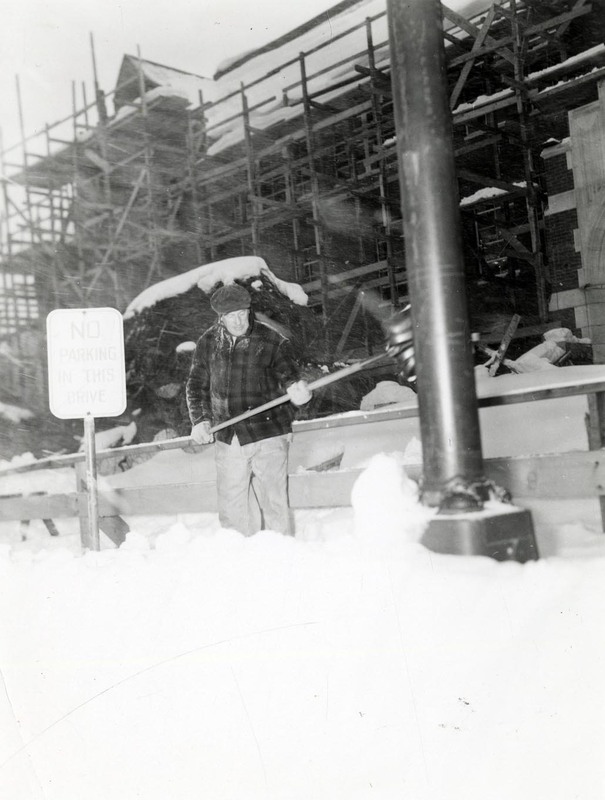 A man shoveling snow next to a sign that reads 'No Parking in this Drive'. A building under construction can be seen in the background.
