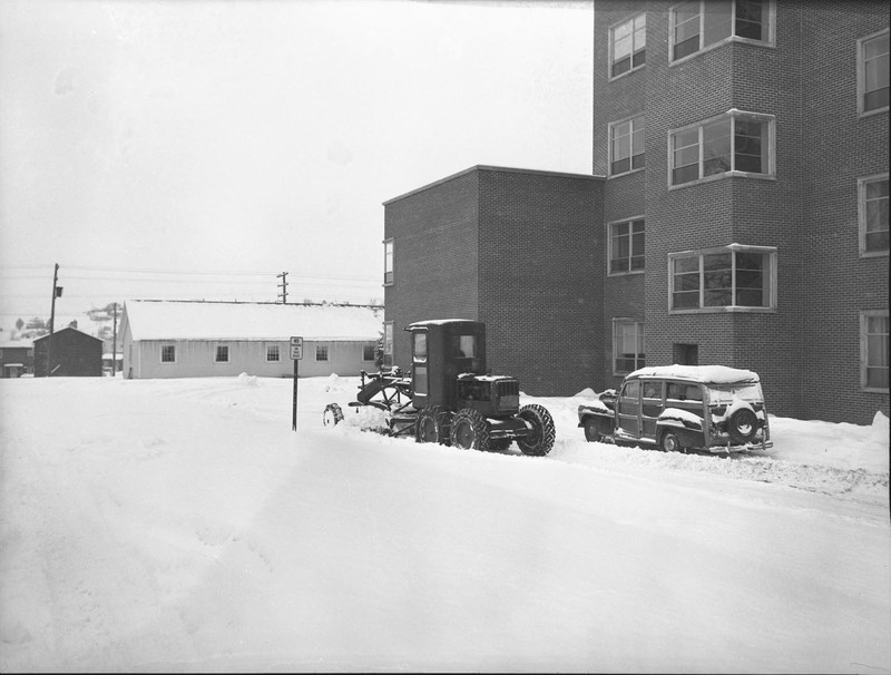 A picture of farming equipment equipped with a snow plow and a automobile outside of the Agricultural Science Building during winter.