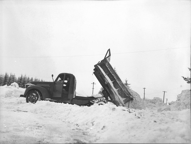 A dump truck dumping snow, the driver can be seen with the truck door open.