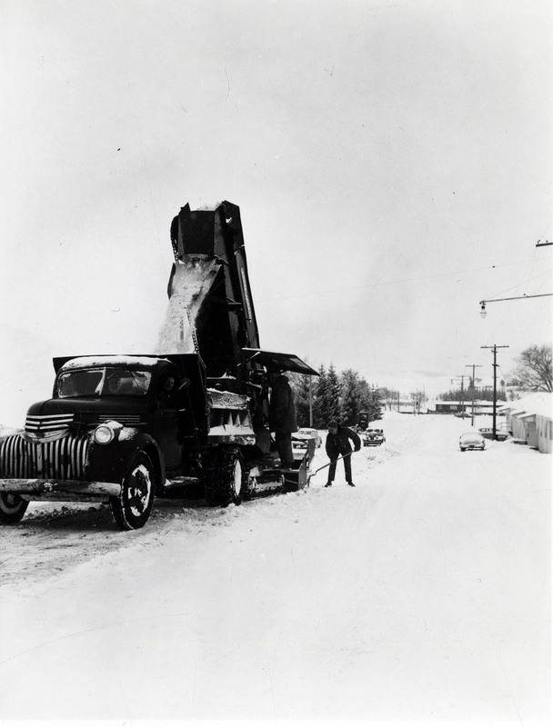 A snow excavator pouring snow into a dump truck during a snowstorm, three men can be seen operating the truck and excavator.