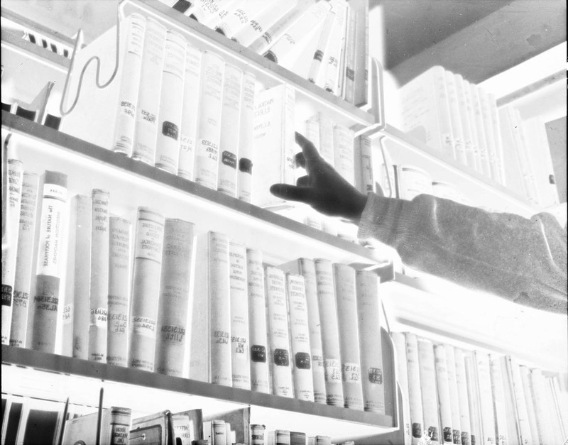 A negative of a photograph of an unidentified person grabbing a book of the shelf in the University of Idaho Library.