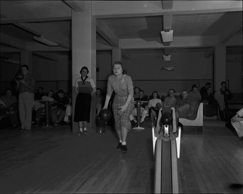 A photograph of a female student mid-bowl in the Student Union Building bowling alley. Large groups of students can be seen in the background.
