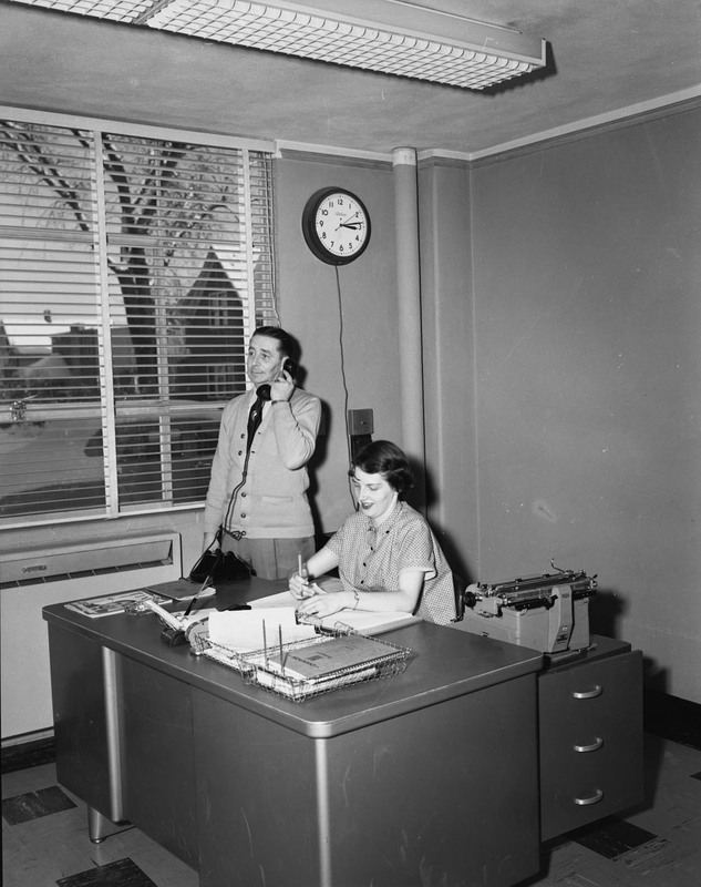 A woman sits behind a desk and writes while a man stands next to her and holds a telephone receiver.