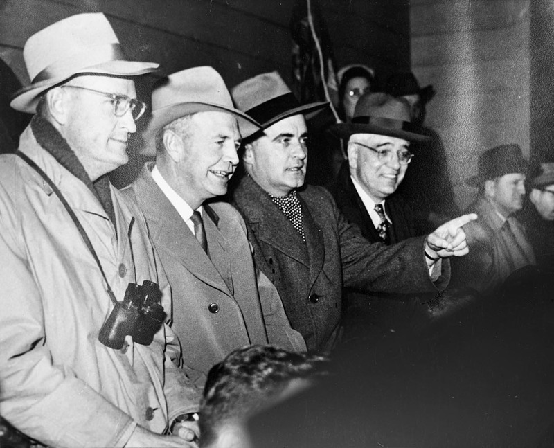 A group of men including President J.E. Buchanan (on the left) are seated in a crowd. One of the men points to the right and Buchanan has a pair of binoculars around his neck.
