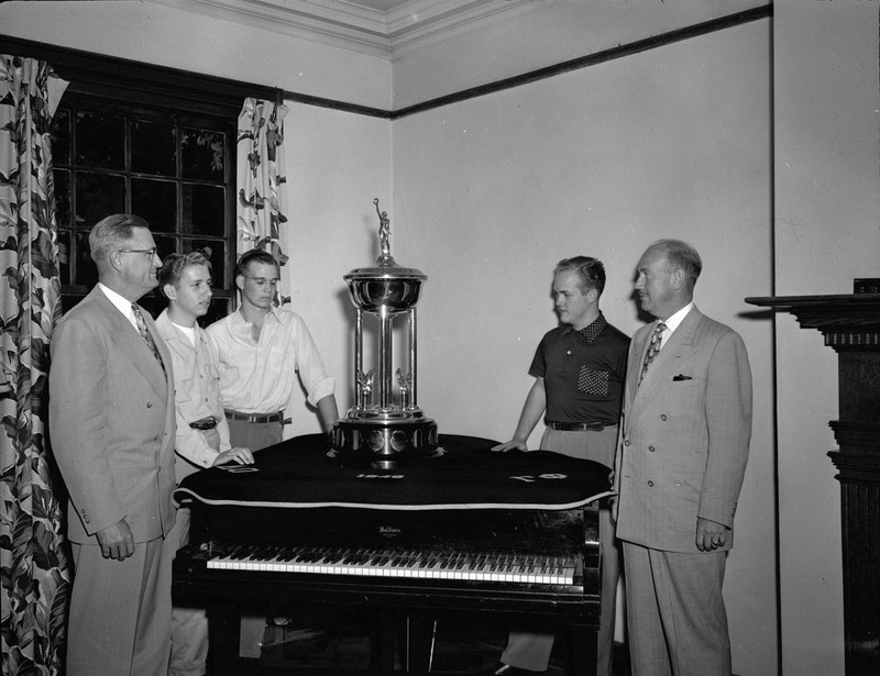 A group of men stand around the Sigma Chi scholarship trophy, which is on top of a piano. Pictured left to right: President Jesse Buchanan, Stanley Swanson, William Mahlik, Richard Coulter, Carl P. Clare.