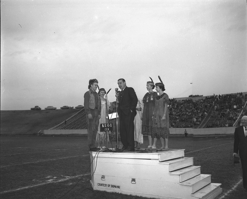 Governor Smylie at the Homecoming game, speaking surrounded costumed students in the football stadium.