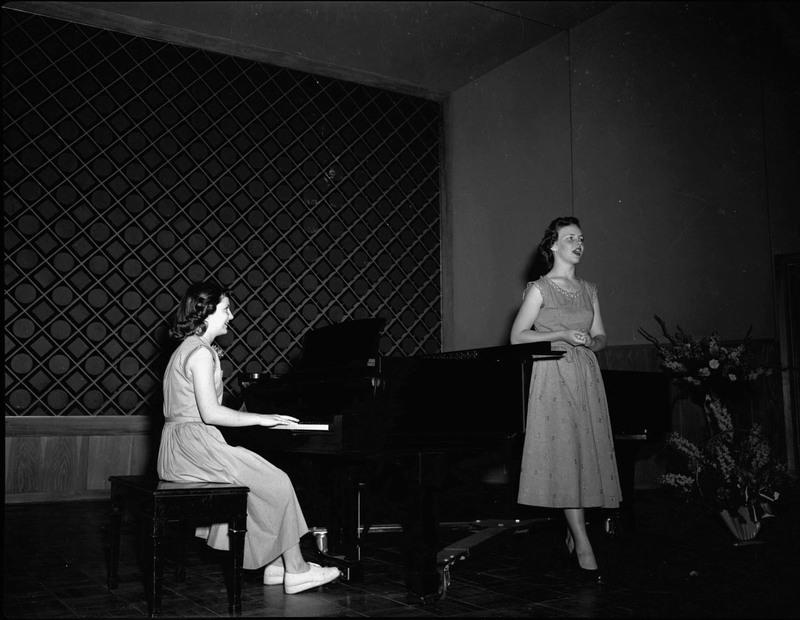 Two women perform, one playing piano and the other singing.
