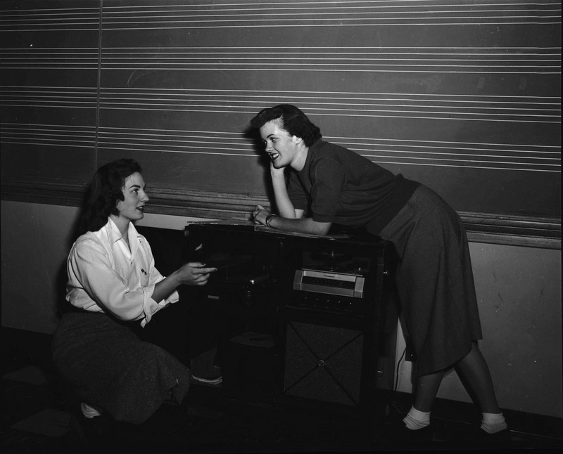 Two unidentified women pose near a media cart. The woman on the left is holding a vinyl record.