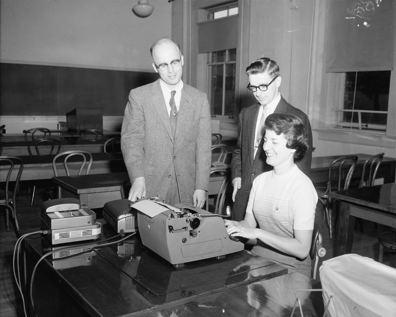 Louise Tatko Cummins, Gem editor, sits at her typewriter with John Hughes and an unidentified man.