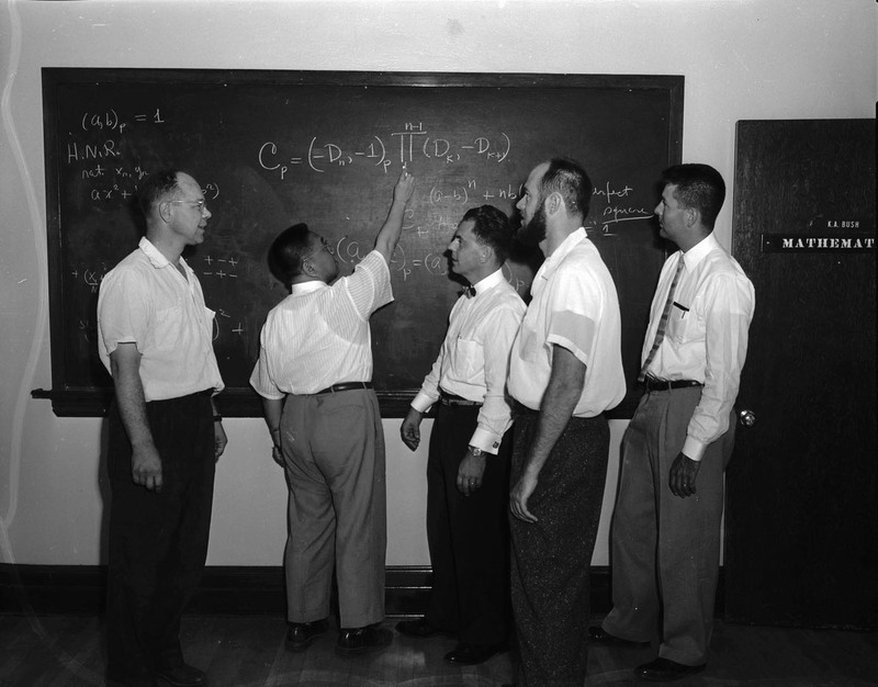 Dr. K. A. Bush writing on a chalkboard while visiting mathematicians from various fields.