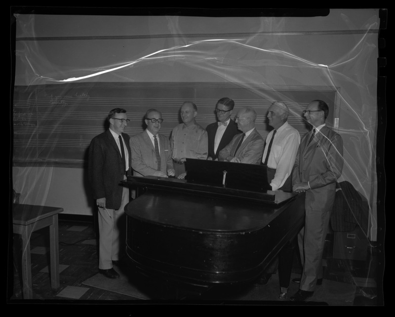 A group of regional singing teachers stand around a piano in a University of Idaho music classroom, Glen Lockery is seen on the far left.