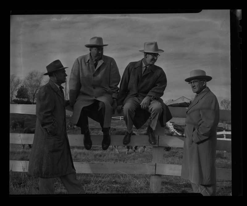 The members of the Idaho Cattlemen's Association dressed in coats and hats outside a fence, two of the men are sitting on the fence. Cows can be seen in the background.