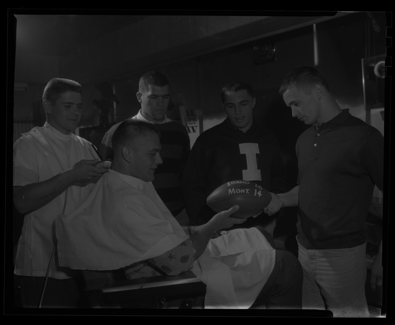 Jim Decko gets his hair cut at the Moscow Barbershop while team members (l-r) Dick Mooney, Mike Mosolf, and Gary Gagnon look at a football with the scores written "Idaho 16, Mont. 14", celebrating Idaho's win over Montana in college football on November 18, 1961 in Old Bronco Stadium, Boise.