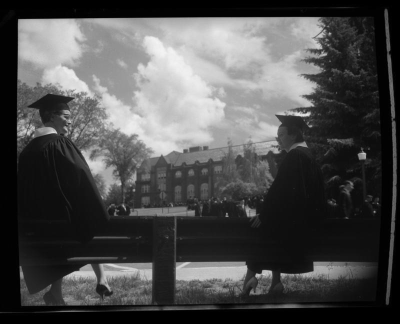 Two graduating senior women sitting in their caps and gowns on a bench during Commencement outside the Administration Building.