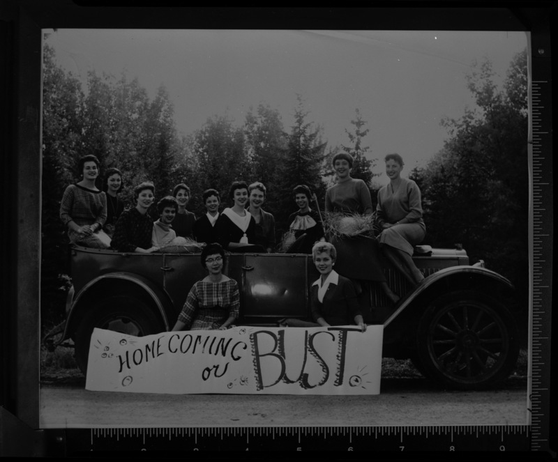 The 13 finalists for homecoming queen pose in a vehicle with a banner that reads "Homecoming or Bust". From left to right: Mary Tsudaka (seated), Charmaine Deitz (seated), Joann Reese, Tonia Peterson, Patt Crowell, Pat Iverson, Kris Bengston, Sharon Matheney, Karla Klamper, Hazel Hunt, Ann Redford, Sandy Wright, and Ann Holden.