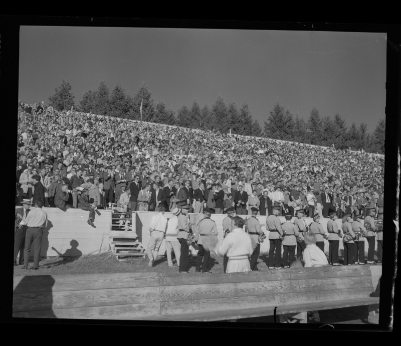 A photograph of filled stands in the football stadium during the homecoming game. The marching band is on the sidelines waiting to perform.