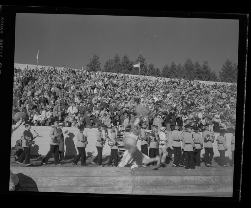 A photograph of filled stands in the football stadium during the homecoming game. The marching band is on the sidelines waiting to perform while a cheerleader jumps in the air in the foreground.