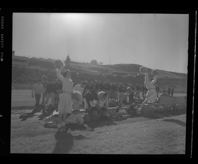Cheerleaders perform on the sidelines, the football team visible in the background.