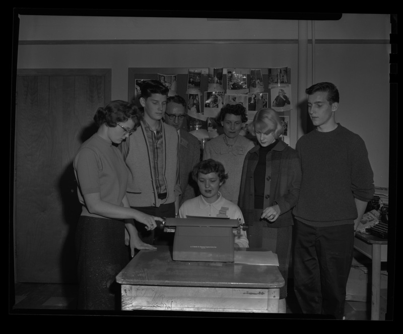Students stand around Delores Hanson at her type writer during a journalism conference at the University of Idaho. Dr. Granville Price, professor of journalism, and Mrs. Stuart observe from behind.