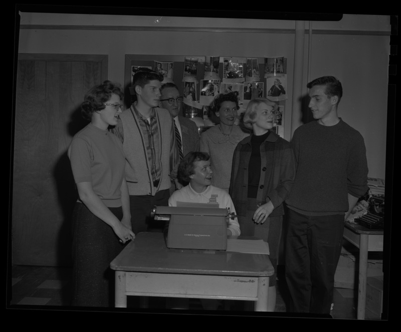 Students stand around Delores Hanson at her type writer during a journalism conference at the University of Idaho. Dr. Granville Price, professor of journalism, and Mrs. Stuart observe from behind.