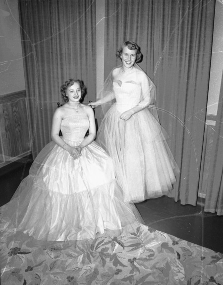 Two women, one sitting, one standing, posing in white gowns.