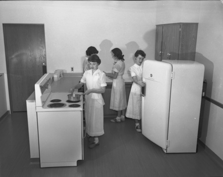 Four women in the Home Economics kitchen, preparing a meal. One is stirring a pot on the stove top, another is looking in the refrigerator, and the other two are in the background.