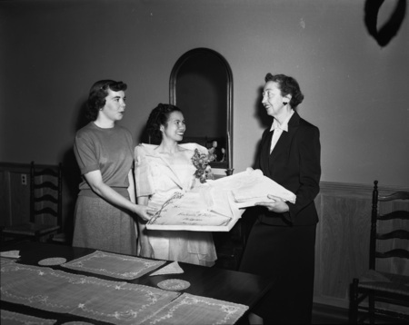 Foreign student from the Philippines, Getrude Vallejos (middle), making a donation to the Department of Home Economics in the company of Margret Ritchie (right), Professor of Home Economics at the University of Idaho, and an unidentified student (left) at a dining table. Vallejos is wearing a white gown and floral boutonniere. A certificate of the donation is in the hands of the woman on the left.