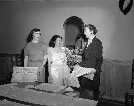Foreign student from the Philippines, Getrude Vallejos (middle), making a donation to the Department of Home Economics in the company of an unidentified faculty member and an unidentified student (left) at a dining table. Vallejos is wearing a white gown and floral boutonniere. A certificate of the donation is in the hands of the woman on the left.