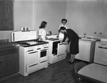 Three women standing around an oven, one opening it.