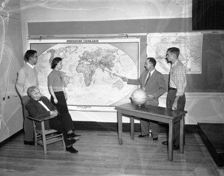 A professor of geography pointing to Syria on a German world map in a classroom. Students are on either side of the map. A globe sits on the desk to the right.