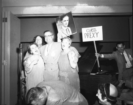 J. E. Buchanan, University of Idaho President, speaking at the class of 1927 reunion with two women on either side of him. A sign saying "class prexy" and a 1927 class portrait of J. E. Buchanan are being held in the background by two men. People can be seen in the foreground.